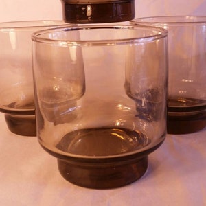 May include: A set of four short drinking glasses made of translucent smoke-colored glass. Each glass features a cylindrical body with a slightly wider, solid base that is darker in tone than the upper portion of the vessel, creating a uniform look.