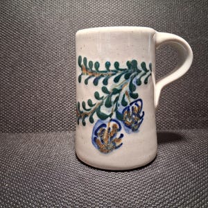 May include: A ceramic mug with a white base and a curved handle. The mug is decorated with a green, blue, and gold floral design. The mug is cylindrical and appears to be handmade.