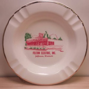 May include: White ceramic ashtray with a gold rim. It features a red and green illustration of a building with the text "Felton Electric, Inc. Jefferson, Wisconsin".