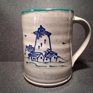May include: A ceramic mug with a white base and a teal rim. The mug features a hand-drawn illustration of a lighthouse and small houses in blue and green. The handle is white and curved.