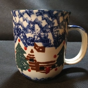 May include: A white ceramic mug with a blue and white speckled glaze. The mug features a winter scene with a brown cabin, two green pine trees, and red ornaments.