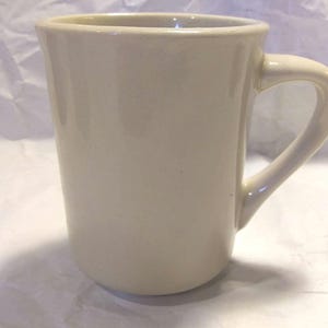 May include: A plain white ceramic mug with a handle. The mug is tall and has a rounded shape.