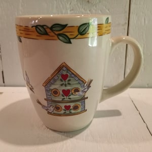 May include: A white ceramic mug with a yellow, green, and blue birdhouse design. The birdhouse has three compartments with red hearts and two birds are flying around it.