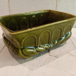 Puede incluir: Una jardinera rectangular de cerámica verde con acabado brillante. La jardinera presenta un diseño arqueado a los lados y un detalle decorativo en forma de cuerda en la base. La jardinera mide aproximadamente 25 cm de largo.