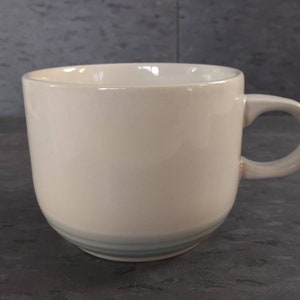 May include: A cream-colored ceramic mug with a rounded body and a curved handle. The mug features a subtle, light blue stripe detail near the base. The mug is designed for hot beverages.