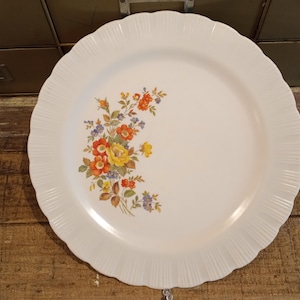 Puede incluir: Un plato blanco con borde festoneado y diseño floral. Las flores son amarillas, naranjas y moradas. El plato tiene una textura acanalada alrededor del borde.