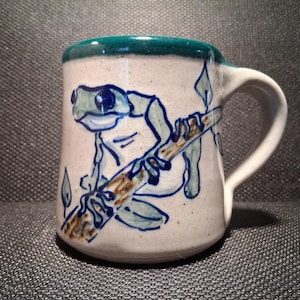 May include: A ceramic mug with a white base and a teal green rim. The mug features a hand-drawn illustration of a frog holding onto a branch. The frog is blue and white, and the branch is brown. The mug has a white handle.