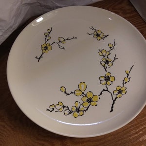 May include: A white ceramic plate with a black and yellow floral design. The flowers are stylized and resemble cherry blossoms.
