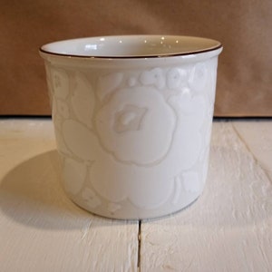 May include: White ceramic mug with a raised floral design. The mug has a brown rim.