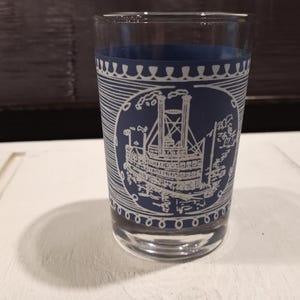May include: A clear glass tumbler with a navy blue band at the top and a white illustration of a steamboat. The design wraps around the glass, and the glass sits on a white surface.