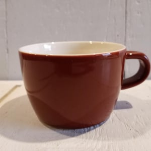 May include: A maroon ceramic coffee mug with a white interior. The mug has a rounded shape and a curved handle. The mug is sitting on a white surface with a wooden background.