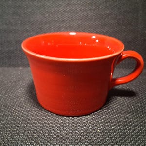 May include: A solid red ceramic coffee cup with a wide, open bowl shape and a single curved handle on the right side. The cup features a smooth, glossy finish and a slightly flared rim, providing a simple and functional design for hot beverages.