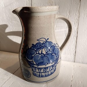 May include: A ceramic pitcher with a blue and white design of apples in a basket. The pitcher has a speckled glaze and a handle.
