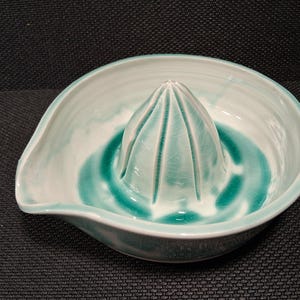 May include: A ceramic citrus juicer with a light teal and white glaze. The juicer has a conical reamer in the center and a spout on the side. The design is a classic kitchen tool for squeezing fresh juice.