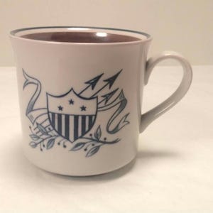 May include: White ceramic mug with a blue and white design of a shield with stars and stripes, arrows, and a ribbon. The mug has a black rim.