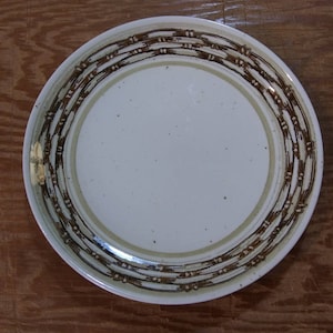 May include: A white ceramic dinner plate with a brown and beige patterned border. The pattern is a series of concentric circles with a textured, woven look.