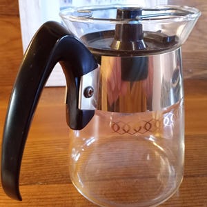 May include: A clear glass coffee pot with a black handle and a silver band around the top. The pot has a decorative pattern on the bottom.