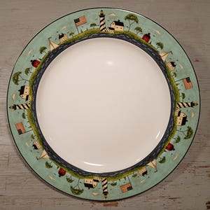 Op de afbeelding: Een wit keramiek dinerbord met een blauwe en groene rand met een herhalend patroon van vuurtorens, zeilboten en Amerikaanse vlaggen.