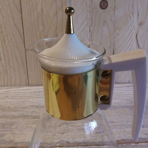 May include: A vintage glass coffee carafe with a white lid and handle. The carafe has a gold-colored metal band around the middle. The lid is topped with a gold-colored finial. The handle is white.