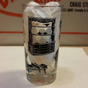 May include: A clear glass with a black and white illustration of a hutch with dishes and a table. The glass is sitting on a white surface.