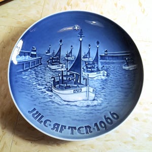 May include: Blue and white decorative plate featuring a harbor scene with several boats. The plate has a nautical theme with the text "Jule Aften 1966" around the edge. The plate is round and has a glossy finish.