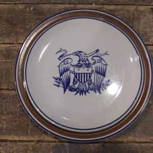 May include: A white ceramic plate with a brown rim features a blue and white illustration of an eagle with a shield. The eagle is holding arrows in its talons and has a banner above its head.