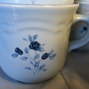 May include: White ceramic mug with a blue floral design featuring a vine with leaves and blue roses. The mug has a handle and a rounded shape.