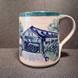 May include: A ceramic mug with a white base, featuring a hand-painted blue and green bridge design. The rim is teal, and the handle is white. This unique mug is perfect for coffee, tea, or any favorite beverage.