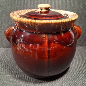 May include: A brown ceramic bean pot with a lid. The pot has two handles and a glossy finish. The lid is a darker shade of brown with a small knob. The rim of the pot and lid have a lighter brown trim.