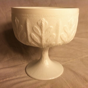 May include: White milk glass pedestal bowl with a leaf design. The bowl has a wide, round base and a tall, slender stem.