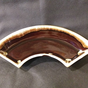May include: A fan-shaped, ceramic serving dish with a glossy, dark brown interior. The rim has a textured, light brown band, and the outer edge is white. The dish has a smooth, curved design, suitable for serving appetizers or snacks.