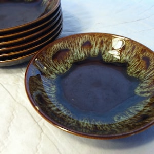 May include: A stack of six brown and blue ceramic plates with a unique glaze. The plates have a dark brown center with a blue hue and a textured, lighter brown and green rim. The plates are arranged on a white surface.