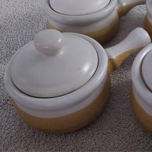 May include: A set of small, white ceramic soup bowls with lids and handles. Each bowl has a band of yellow-tan color around the base. The lids have a small, round knob on top.