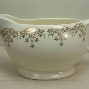 May include: A white ceramic gravy boat with a gold floral design around the rim. The boat has a handle and a rounded base.
