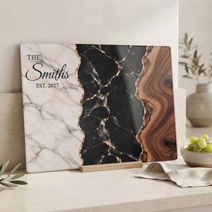May include: A rectangular cutting board featuring a tri-panel design with white marble on the left, black marble in the center, and brown wood grain on the right. The white section displays the black text THE Smiths EST. 2027.
