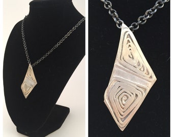 Puede incluir: Un collar de plata con un gran colgante geométrico con un diseño abstracto en espiral. El colgante está unido a una cadena negra.