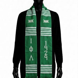 Pode incluir: Um cachecol verde com listras horizontais brancas nas extremidades. O cachecol exibe as letras gregas Phi Beta Sigma verticalmente de um lado e o ano 1914 verticalmente do outro, finalizado com uma bainha de franjas na parte inferior.