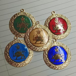 Puede incluir: Cinco colgantes circulares de metal dorado con un borde texturizado y un emblema central de un león con una espada y una corona. Los emblemas están sobre fondos esmaltados en verde, rojo, beige y dos tonos de azul, cada uno con una argolla.
