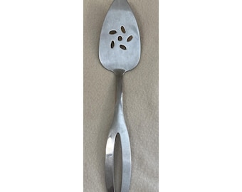 Könnte beinhalten: Ein silberfarbener Tortenheber aus Metall mit einer spitz zulaufenden, dreieckigen Klinge, die ein dekoratives Lochmuster aus fünf kleinen, kreisförmig angeordneten ovalen Öffnungen und einen Griff mit einer großen, länglichen Aussparung aufweist.