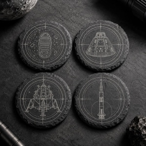 May include: Four round black slate coasters featuring white laser-engraved space exploration designs. The designs include a lunar boot print, a command module, a lunar lander, and a Saturn V rocket, each centered within a circular crosshair pattern.