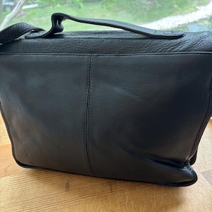 Puede incluir: Bolso mensajero de cuero negro con asa superior y bandolera ajustable. El bolso presenta un cierre de solapa y un dise&ntilde;o cosido. El bolso est&aacute; sobre una superficie de madera.