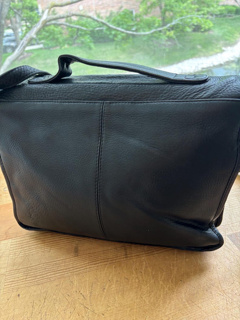 Puede incluir: Bolso mensajero de cuero negro con asa superior y bandolera ajustable. El bolso presenta un cierre de solapa y un dise&ntilde;o cosido. El bolso est&aacute; sobre una superficie de madera.