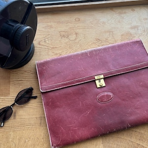 May include: A burgundy leather portfolio with a gold-colored clasp and a small oval emblem. Black sunglasses rest on a wooden surface next to the portfolio. A black object is in the background.