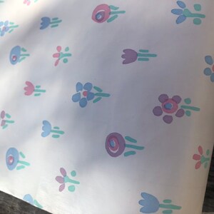 May include: White paper with a repeating pattern of stylized flowers in shades of pink, blue, and purple. The floral design includes various shapes, such as tulips and circular blooms, with green stems and leaves. The paper is slightly creased.