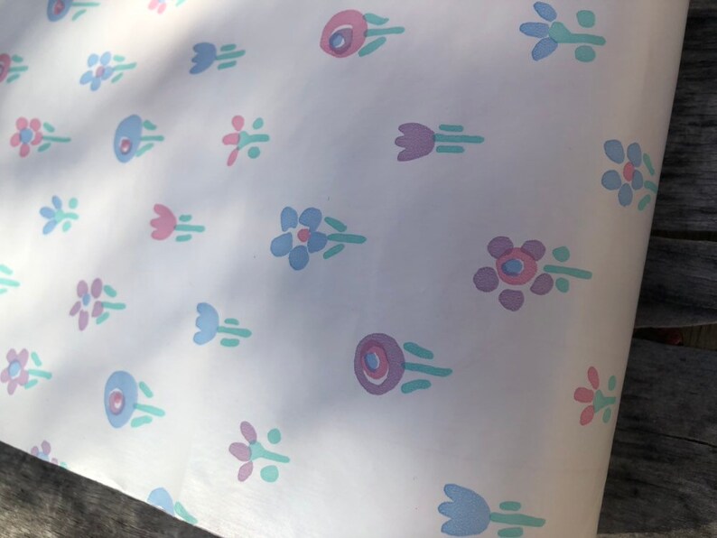 May include: White paper with a repeating pattern of stylized flowers in shades of pink, blue, and purple. The floral design includes various shapes, such as tulips and circular blooms, with green stems and leaves. The paper is slightly creased.