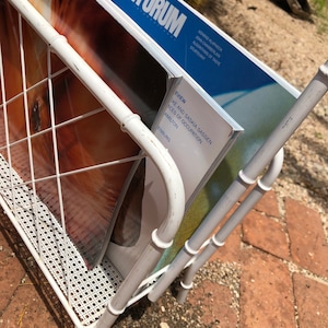 May include: White metal magazine rack with two magazines inside. The top magazine is titled "Artforum" and the bottom magazine is titled "Eview" with the text "Mike and Saskia Sassen, Aces of Occupation, Hamilton, Enburg".