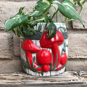 May include: A white ceramic planter with a mushroom design. The planter has three red mushrooms with green leaves and a gray and white striped background. A green plant is growing in the planter.