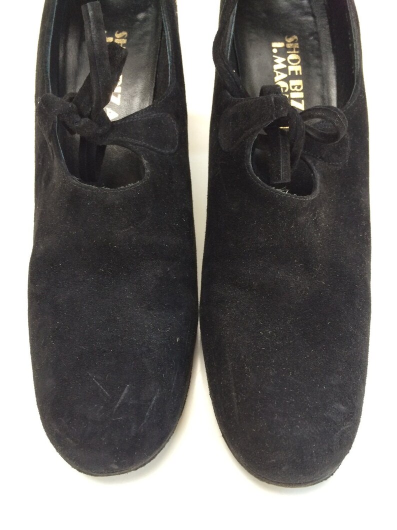 May include: A pair of black suede shoes with a lace-up design. The shoes have a round toe and a low heel. The brand name "Shoe Biz I.Magi" is printed on the inside of the right shoe.