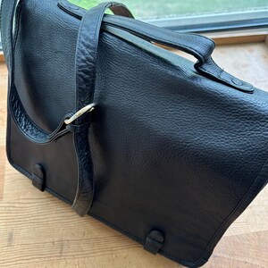 Puede incluir: Un bolso mensajero de cuero negro con una superficie texturizada. El bolso tiene un asa superior, una correa para el hombro y un cierre de solapa con dos hebillas. El bolso est&aacute; sobre una superficie de madera.