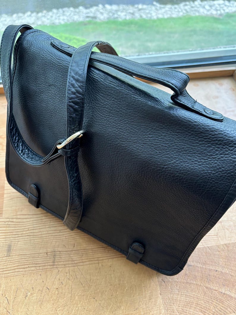 Puede incluir: Un bolso mensajero de cuero negro con una superficie texturizada. El bolso tiene un asa superior, una correa para el hombro y un cierre de solapa con dos hebillas. El bolso est&aacute; sobre una superficie de madera.